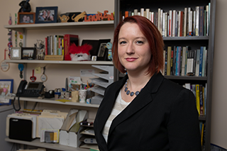 Image shows alumna Lauren Cagle standing and smiling at the camera in her office in front of a bookshelf filled with books, and shelves with books, a phone, files, picture frames, and various other decorations.