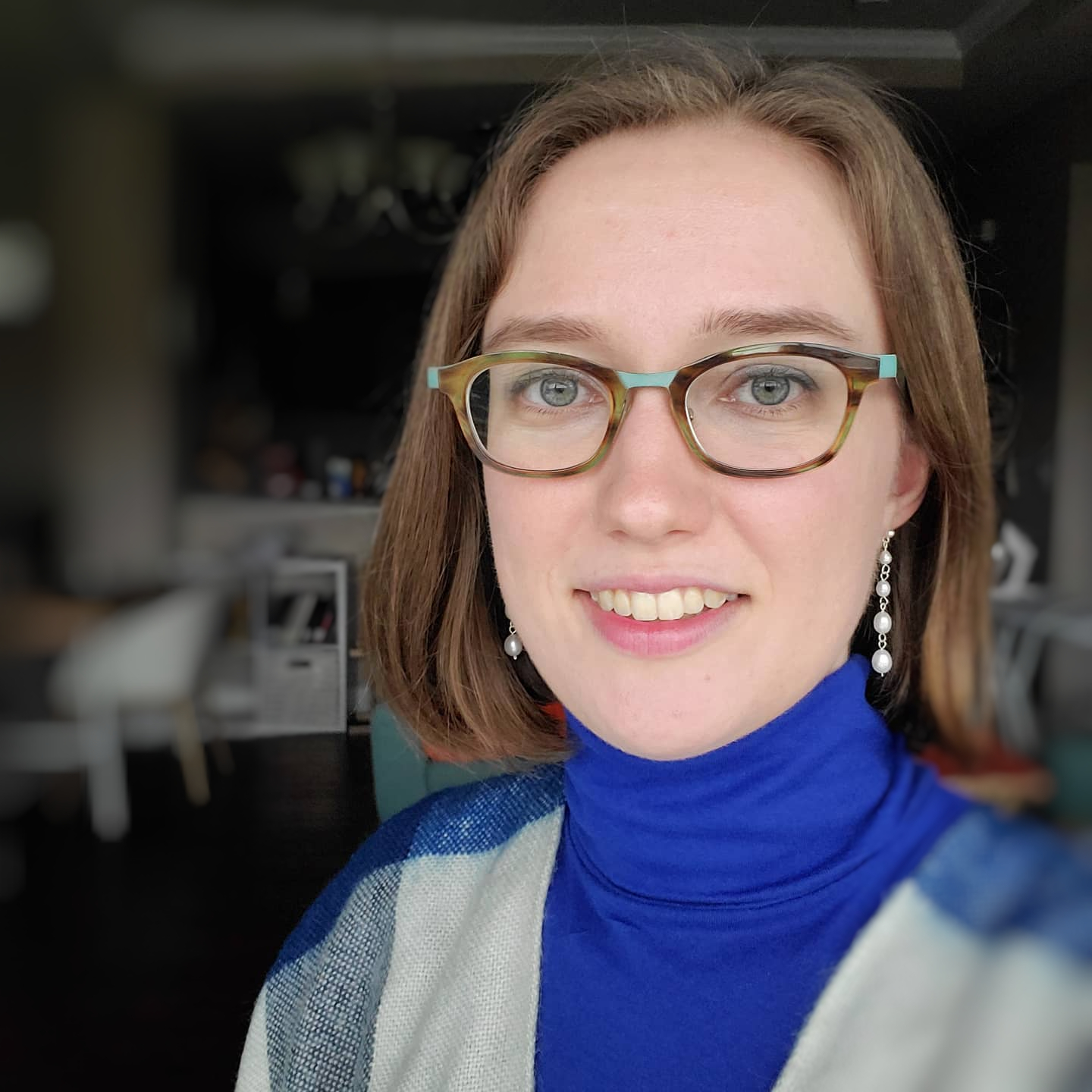 A white woman with brown hair wearing glasses, a blue turtleneck, and a grey and white-checked sweater