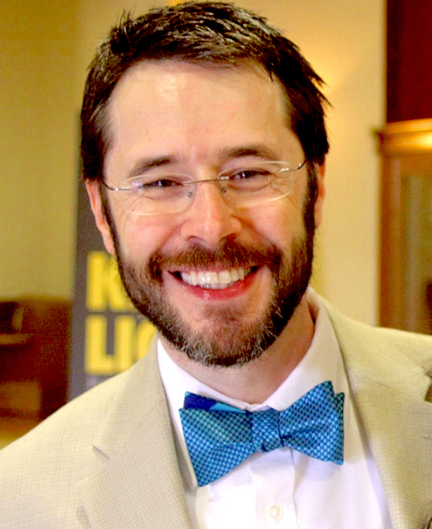 A white man with brown hair and a brown beard, wearing glasses, a white shirt, cream blazer, and blue bowtie