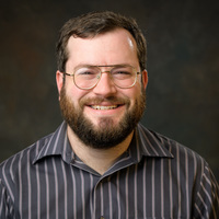 A white man with brown hair and a brown beard, wearing glasses and a black shirt with white. pinstripes