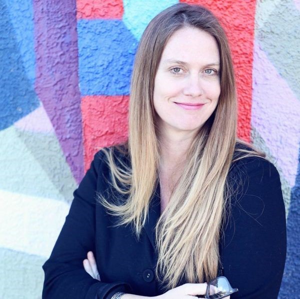 A white woman with long blond hair wearing a navy blazer and standing in front of a multicolored mural