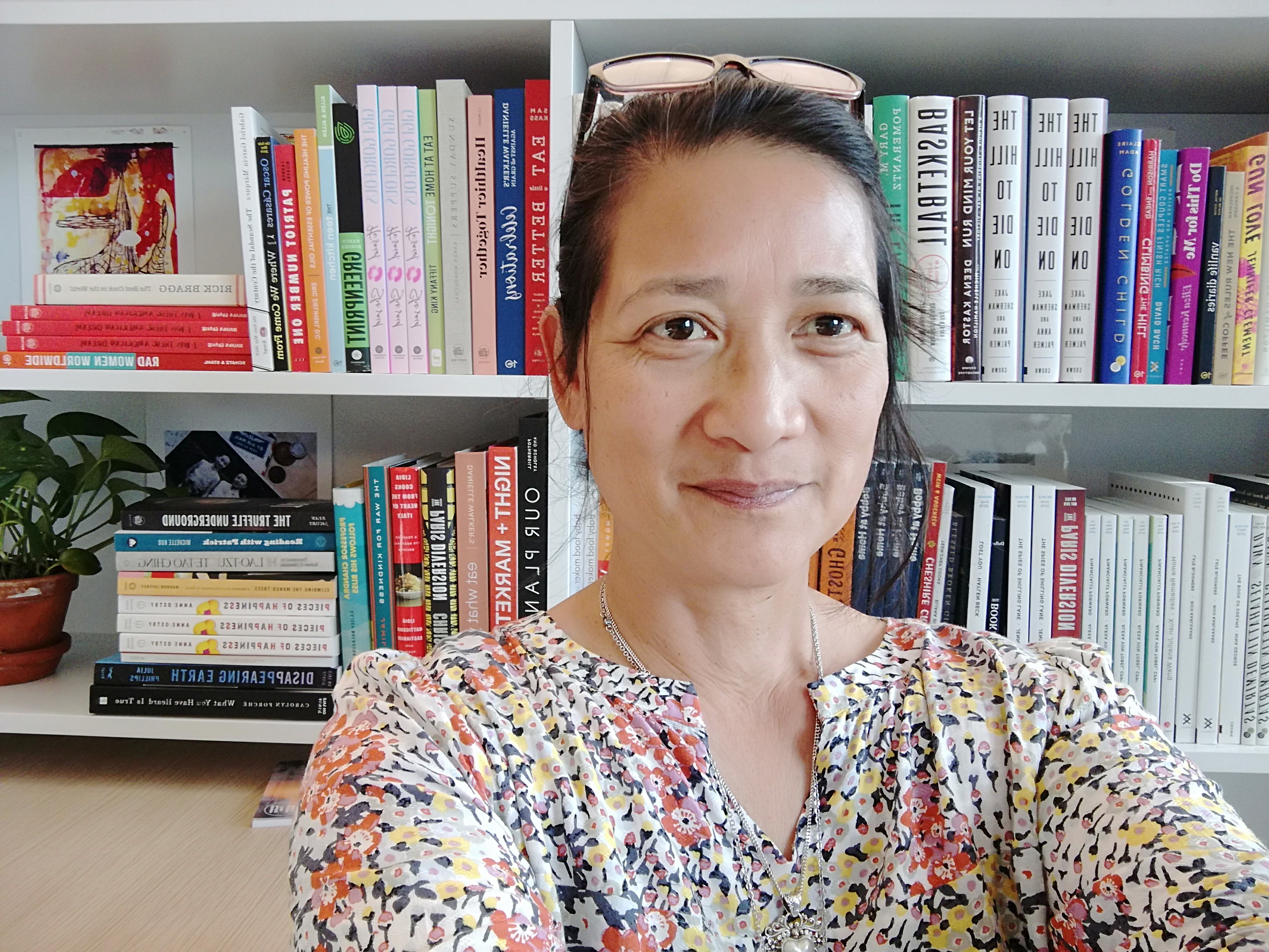 An Asian woman with dark hair, wearing sunglasses on her head, a floral print blouse, and a silver necklace, sitting in front of a bookcase