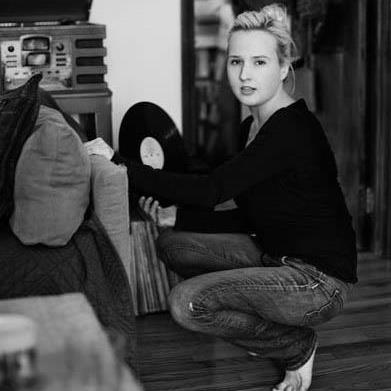Black and white photo of a white woman wearing a black sweater a jeans, crouching and holding a record