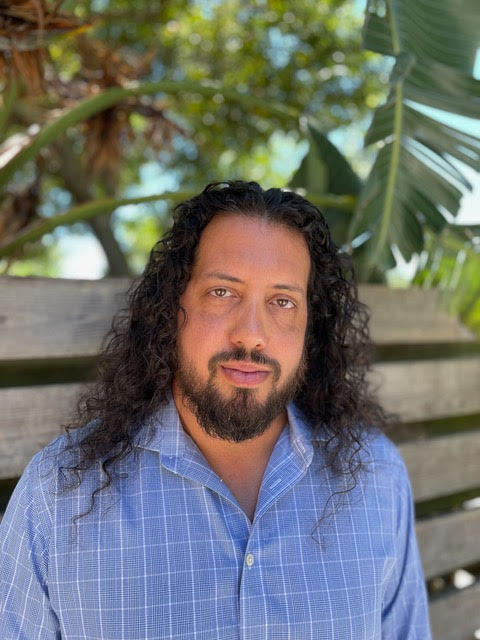 A tanned man with long, dark curly hair and a dark beard wearing a checkered blue shirt