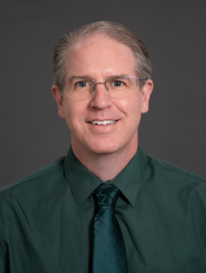 man with green shirt and tie smiling against neurtal dark grey background
