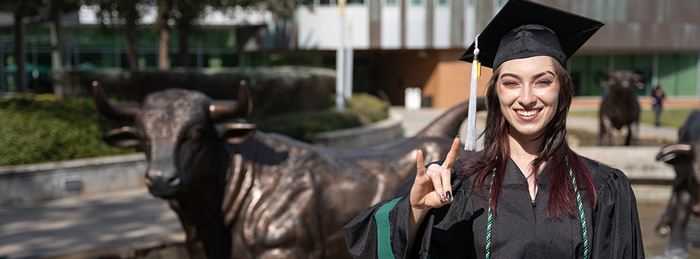 graduate in cap and gown with her hand in bulls sign, standing in front of bronze bull statue