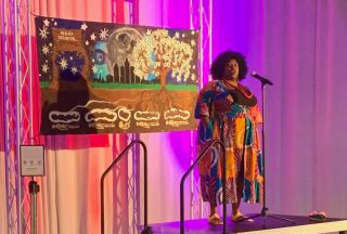 Spoken word poet, Mis Wright, delivering a poem on a small elevated stage with pink/purple lighting and a large painted canvas displayed behind her painted by visual artist Lakeema Williams.
