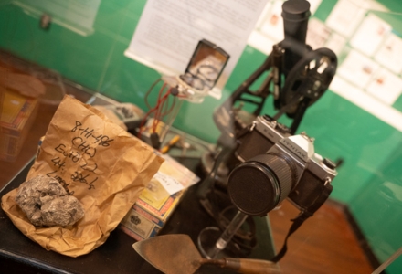 The exhibit includes artifacts in the form of technology used by anthropology faculty over the past 60 years; shown here are a few tools used by archaeologists, including paper bags once used for curation (now replaced by archival quality plastic), a 35 mm SLR camera (now largely replaced by smart phones), and a survey transit (now supplanted by GPS, total stations, and LiDAR, and a trowel (still widely used).  (Photo by Corey Lepak)