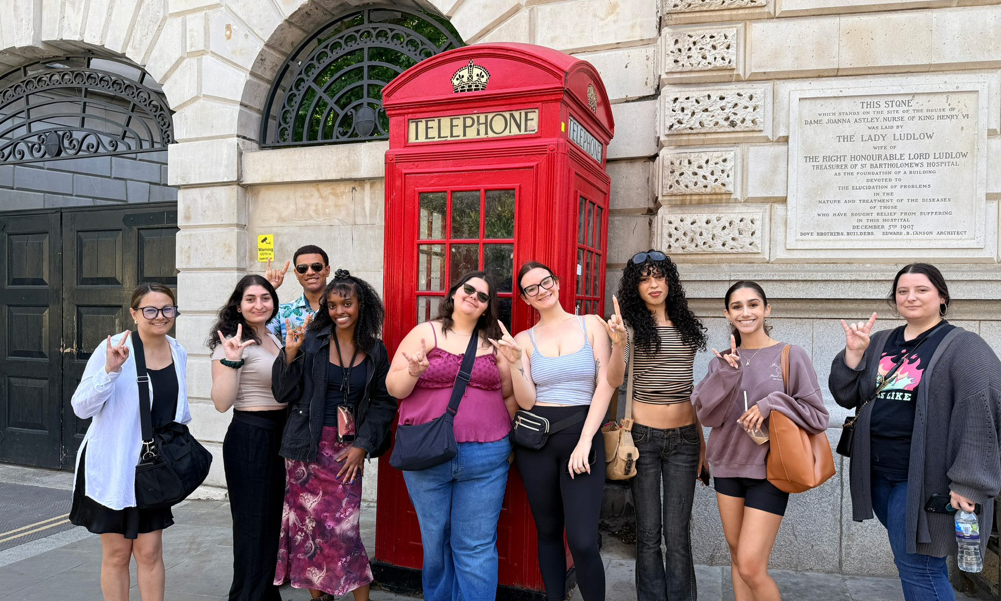 Group of people standing together on a city street beside a classic red British telephone booth, posing in front of a stone building with arched windows and a commemorative plaque.