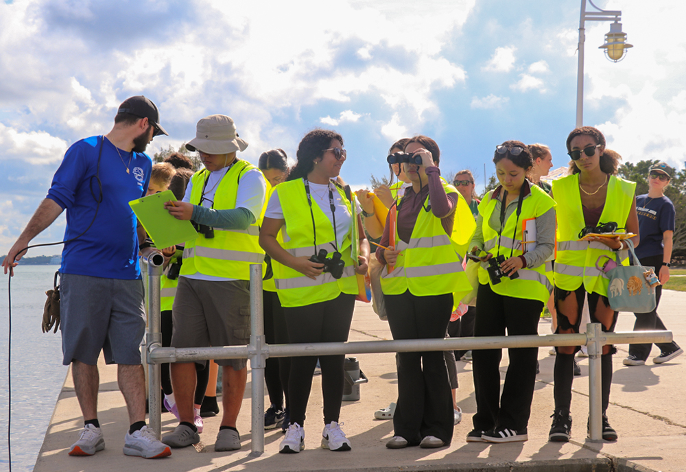 A group of high school students wearing bright yellow safety vests stand on a waterfront pier with cameras and binoculars. A scientist or instructor in a blue shirt holds a clipboard and speaks to them. The group faces the water under a cloudy sky, participating in an outdoor field activity.