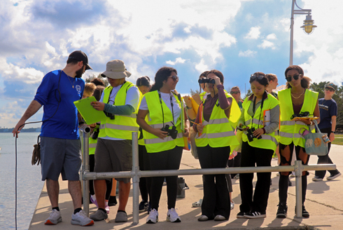 A group of high school students wearing bright yellow safety vests stand on a waterfront pier with cameras and binoculars. A scientist or instructor in a blue shirt holds a clipboard and speaks to them. The group faces the water under a cloudy sky, participating in an outdoor field activity.