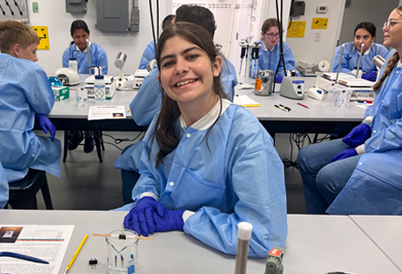 Students wearing blue lab coats and purple gloves work at lab benches with microscopes, pipettes, and sample containers. One student in the foreground smiles while preparing materials for a hands-on science activity.