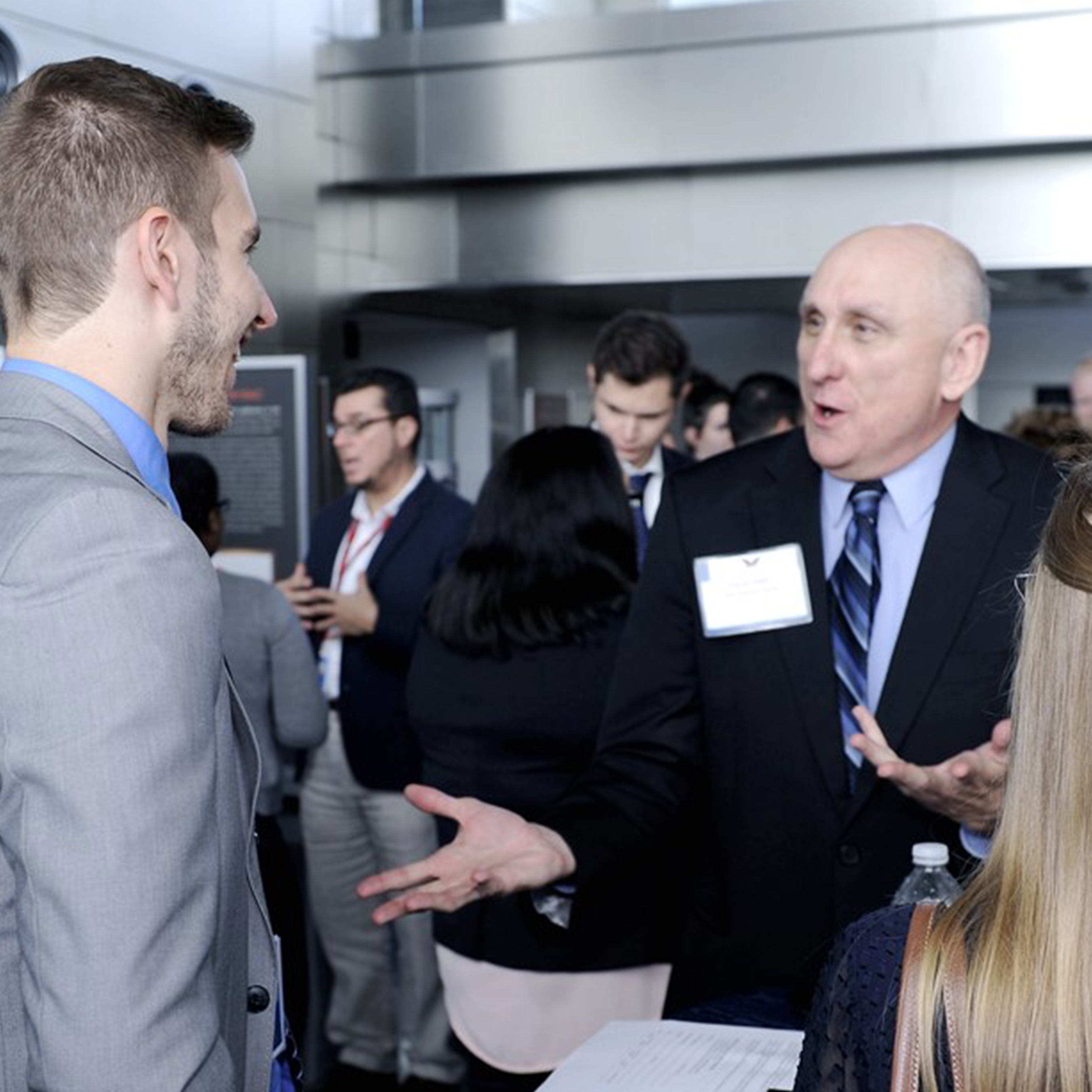 A male DIA intern in a grey suit smiles as they converse with an older DIA agent dressed in a black suit with a blue striped tie.