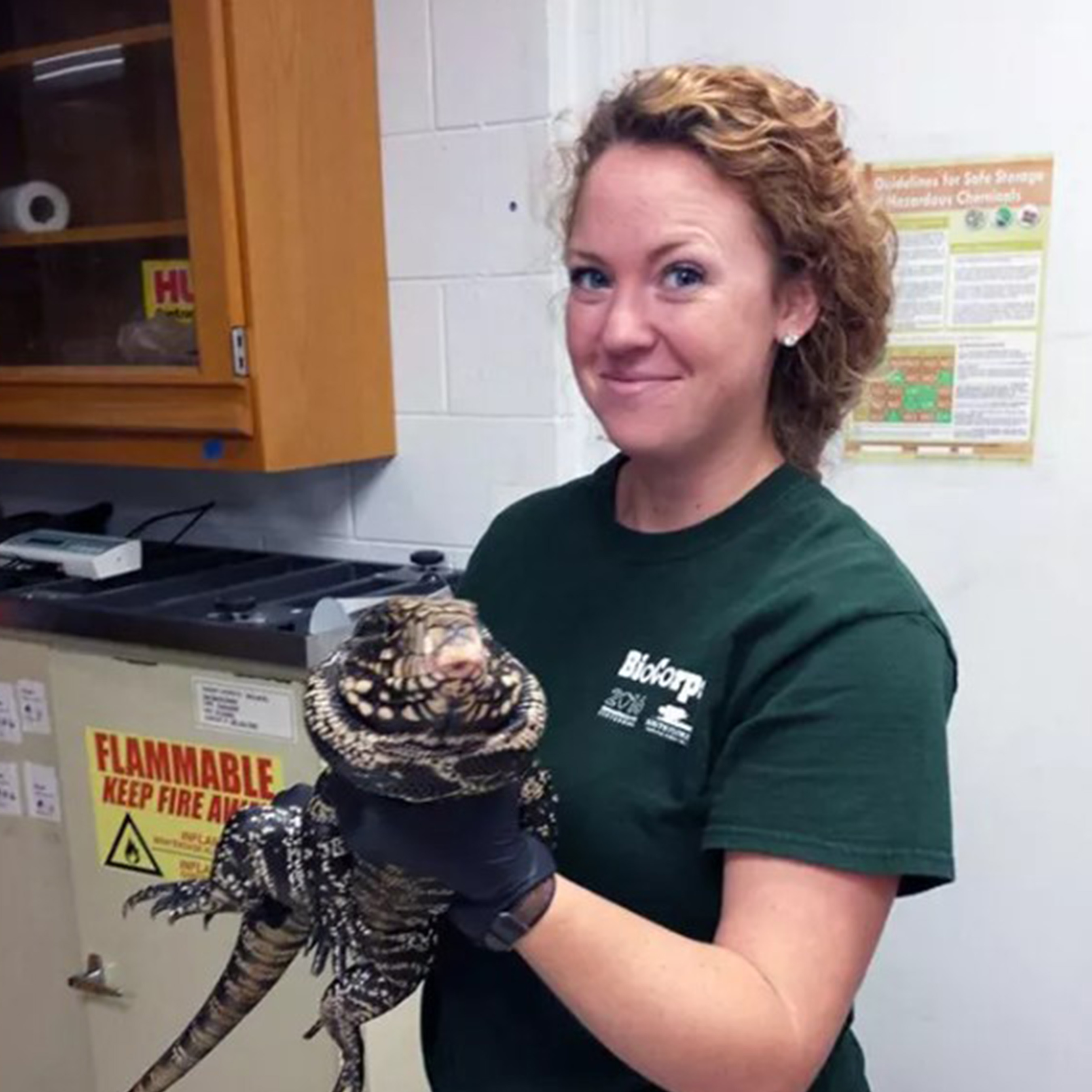 A female Biocorp intern in a green t-shirt holds a large lizard inside of a lab. 