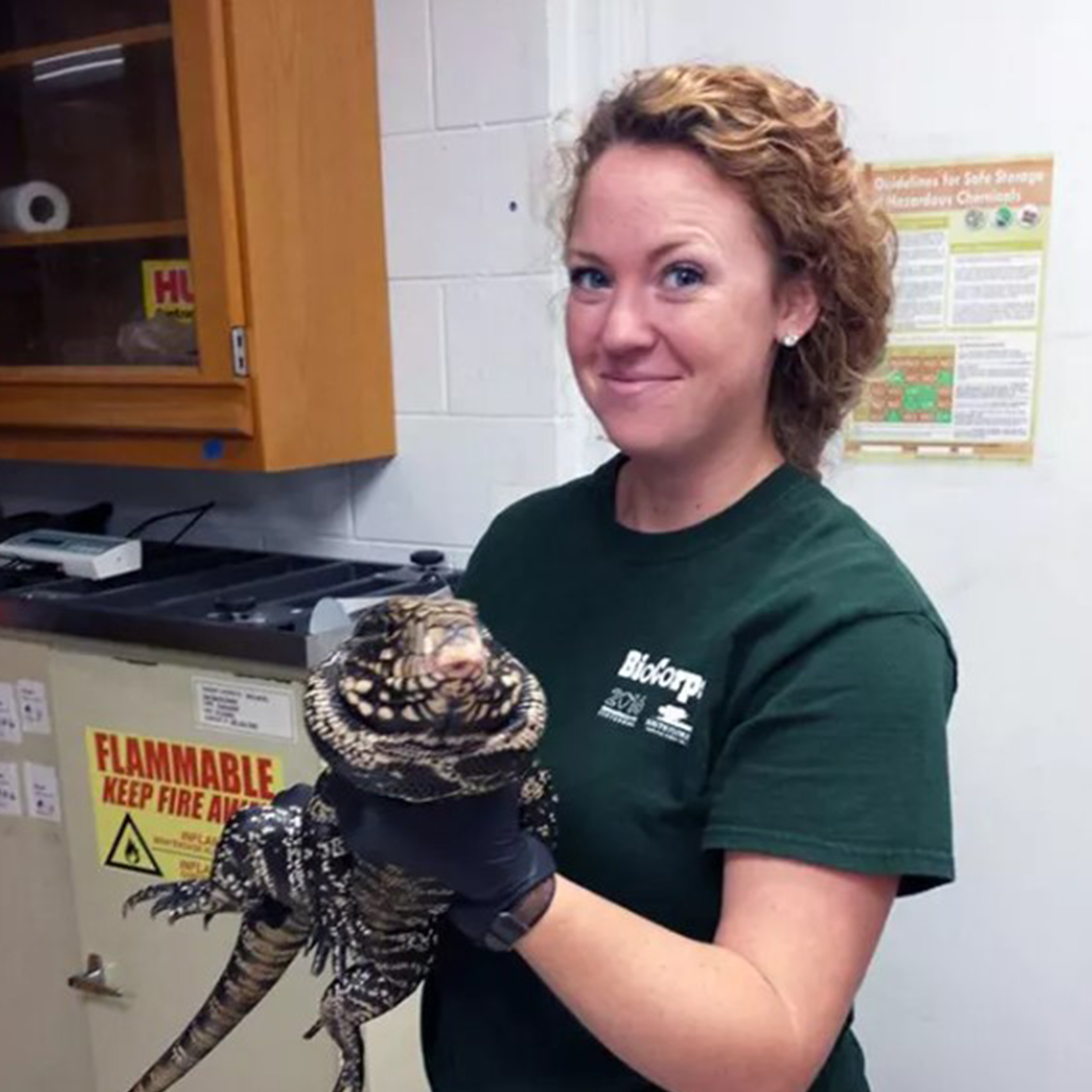 A female Biocorp intern in a green t-shirt holds a large lizard inside of a lab.