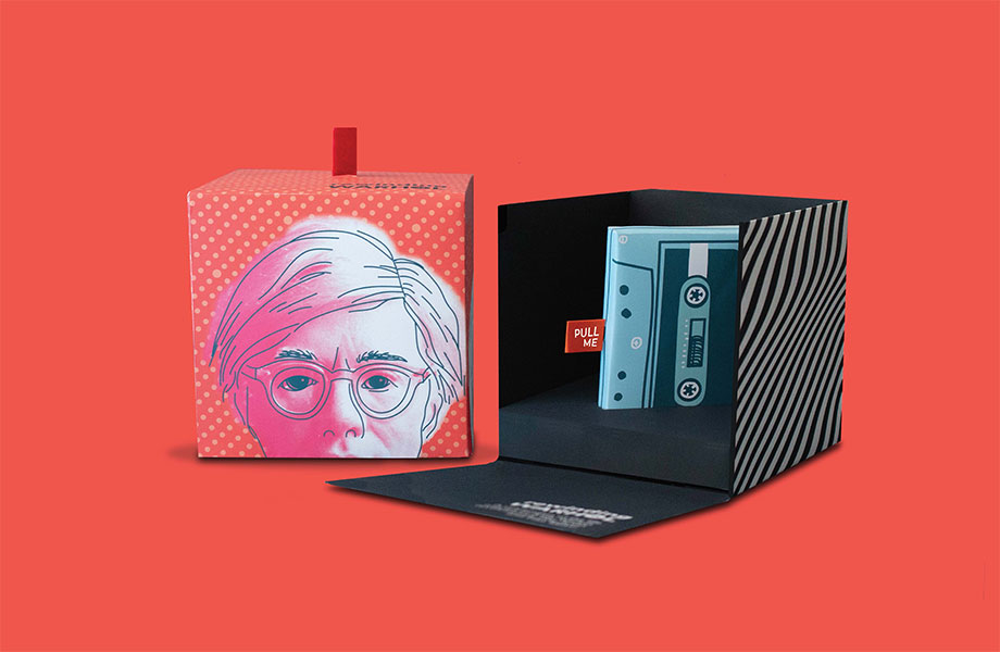 Graphic design work by Tessa Wilson, 2020 graduate. Two boxes sit side by side against a coral orange background. The box on the left is closed, the cover shows a person's face from the nose up. They have short hair and their eyes stare through their glasses. The box on the right has a black and grey zebra print. It sits open, revealing a casette with a red tab sticking out that reads "PULL ME."