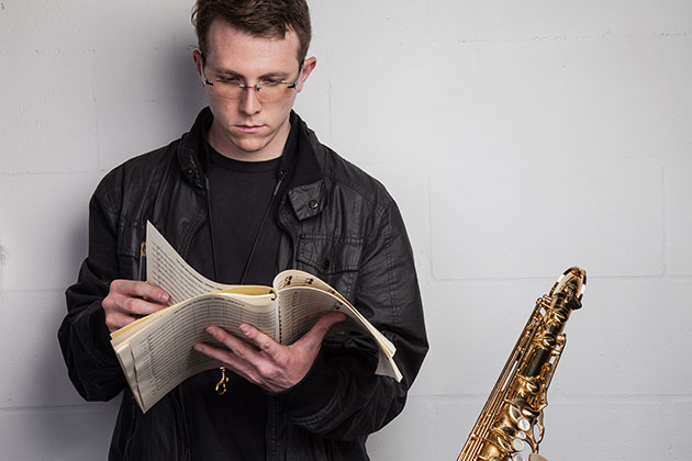 Zachary Bornheimer looks at sheet music in his hands with a saxophone to his left in a studio portait