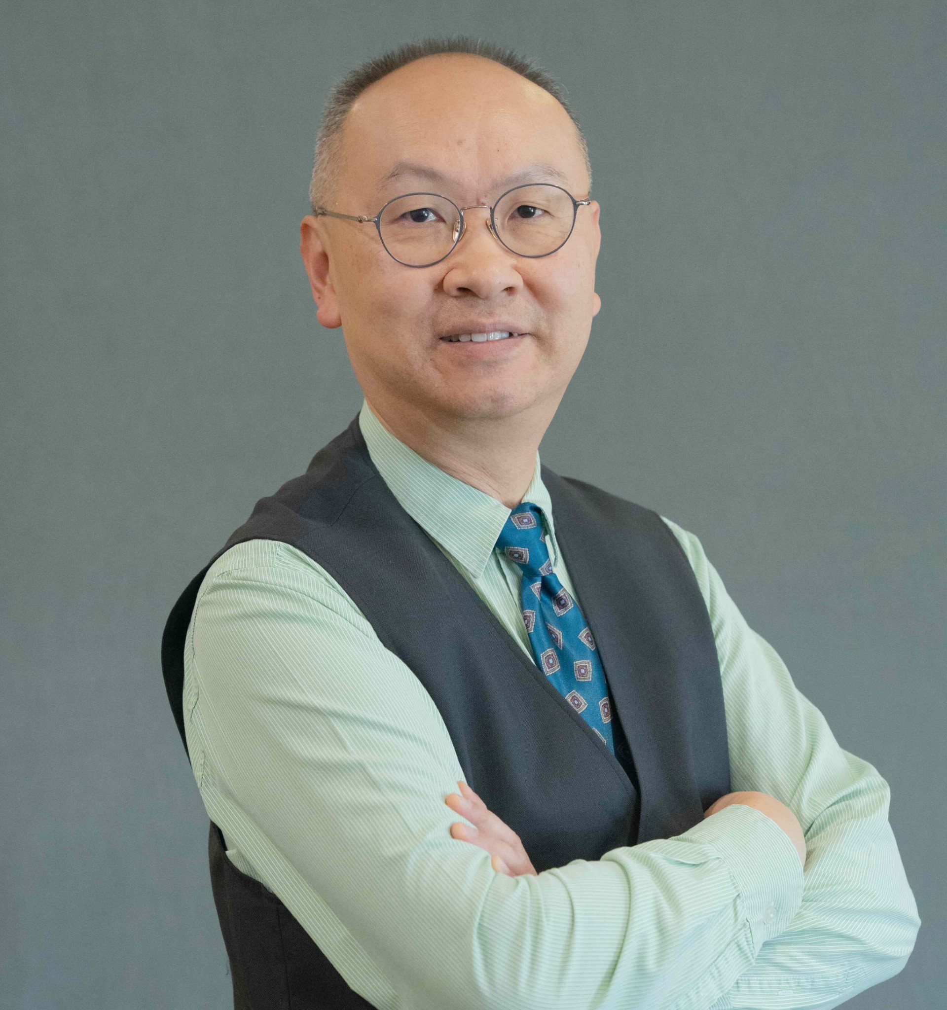Victor Fung wearing a green shirt, blue tie, and grey vest in front of a grey background