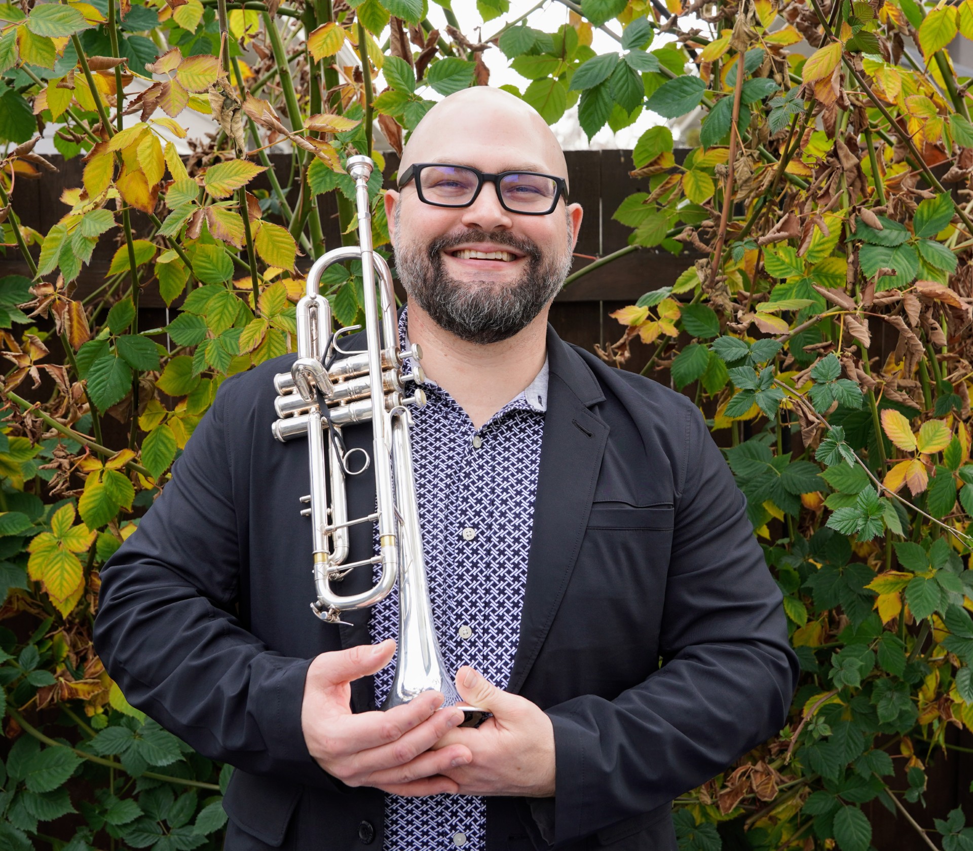 Derek Ganong wearing a blue patterned shirt with a blue blazer holding a trumpet