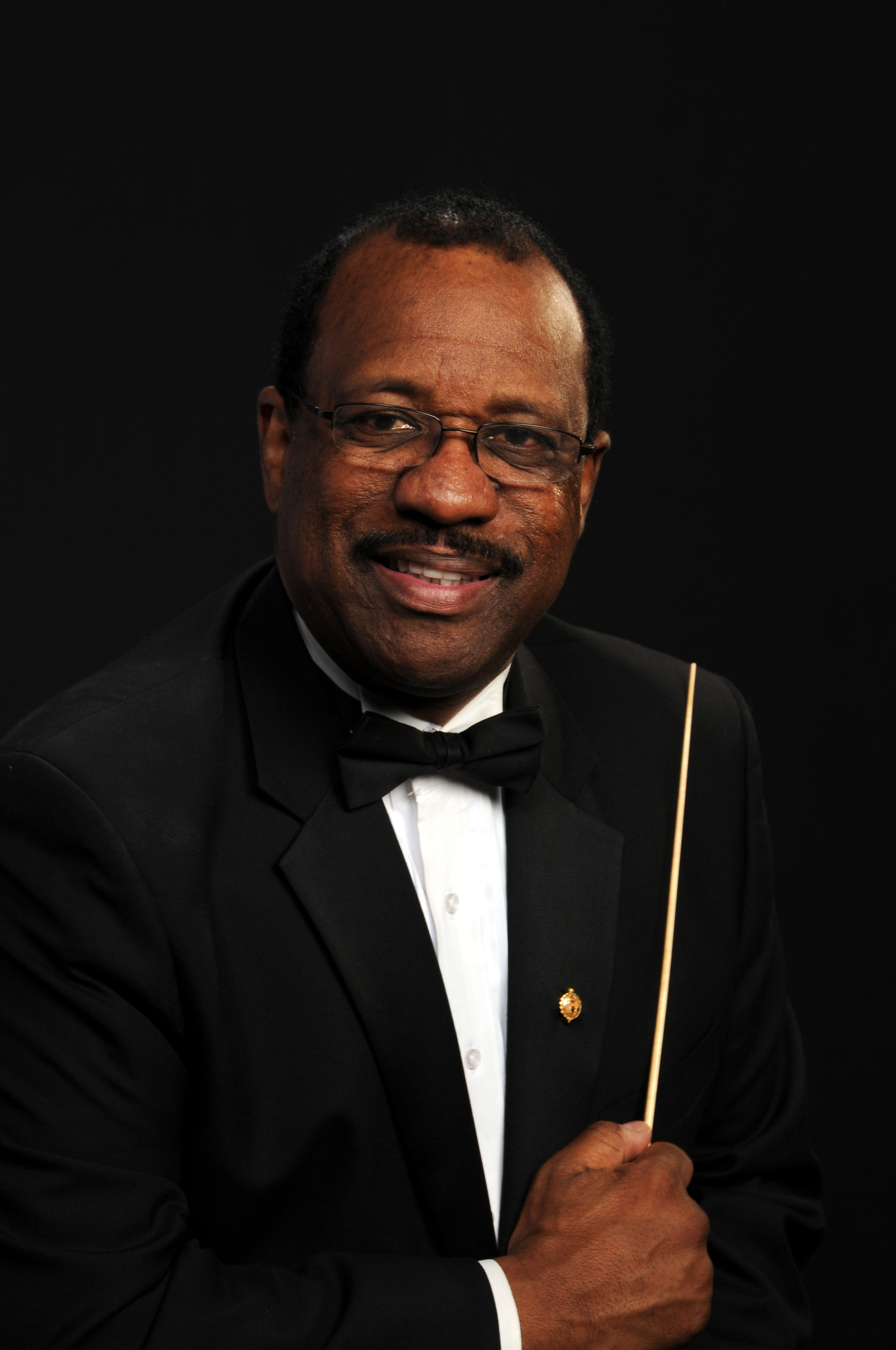 Headshot image of Alfred Watkins wearing a tuxedo jacket and bowtie holding a baton in front of a black background.