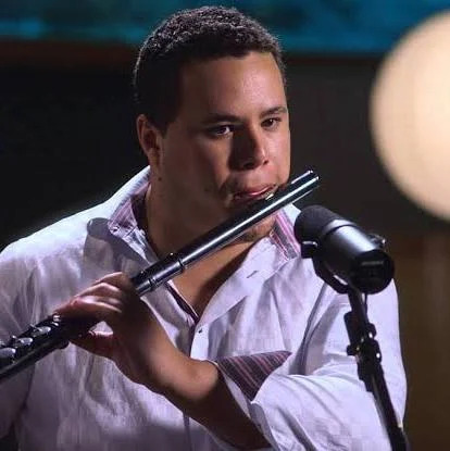Jose Valentino Ruiz wearing a long sleeved collared shirt playing the flute in front of a microphone