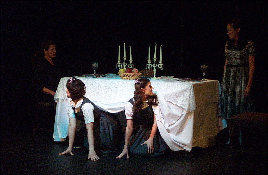A man sits at the end of a dining table while a girl watches him. Two girls crawl out from underneath the tablecloth, one looking at the man and the other looking a the other girl who is watching the man.