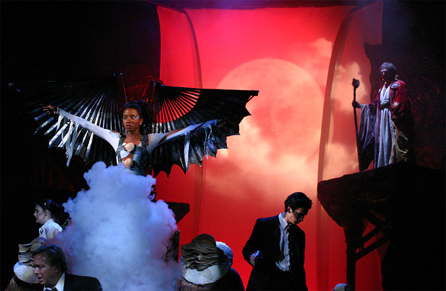A woman with wings stands in a cloud of smoke, being watched by a man with robes decorated with a pair of eyes and holding a staff. Surrounding the woman are characters all turned away from her, some of these characters have warped green faces. 