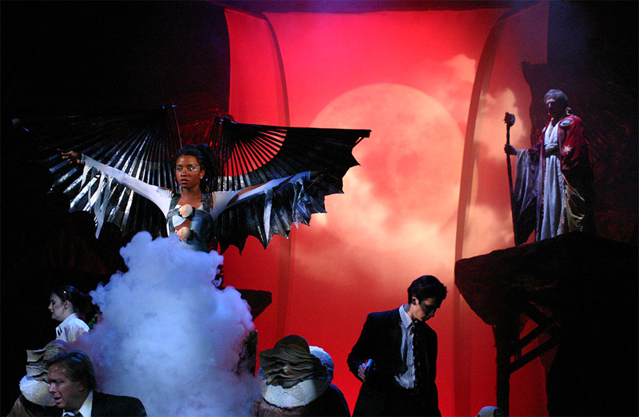 A woman with wings stands in a cloud of smoke, being watched by a man with robes decorated with a pair of eyes and holding a staff. Surrounding the woman are characters all turned away from her, some of these characters have warped green faces.