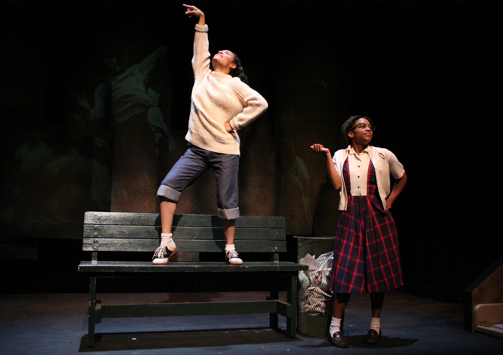 Two women stand in different poses onstage, one standing on top of a bench while the other stands on the ground next to the bench.