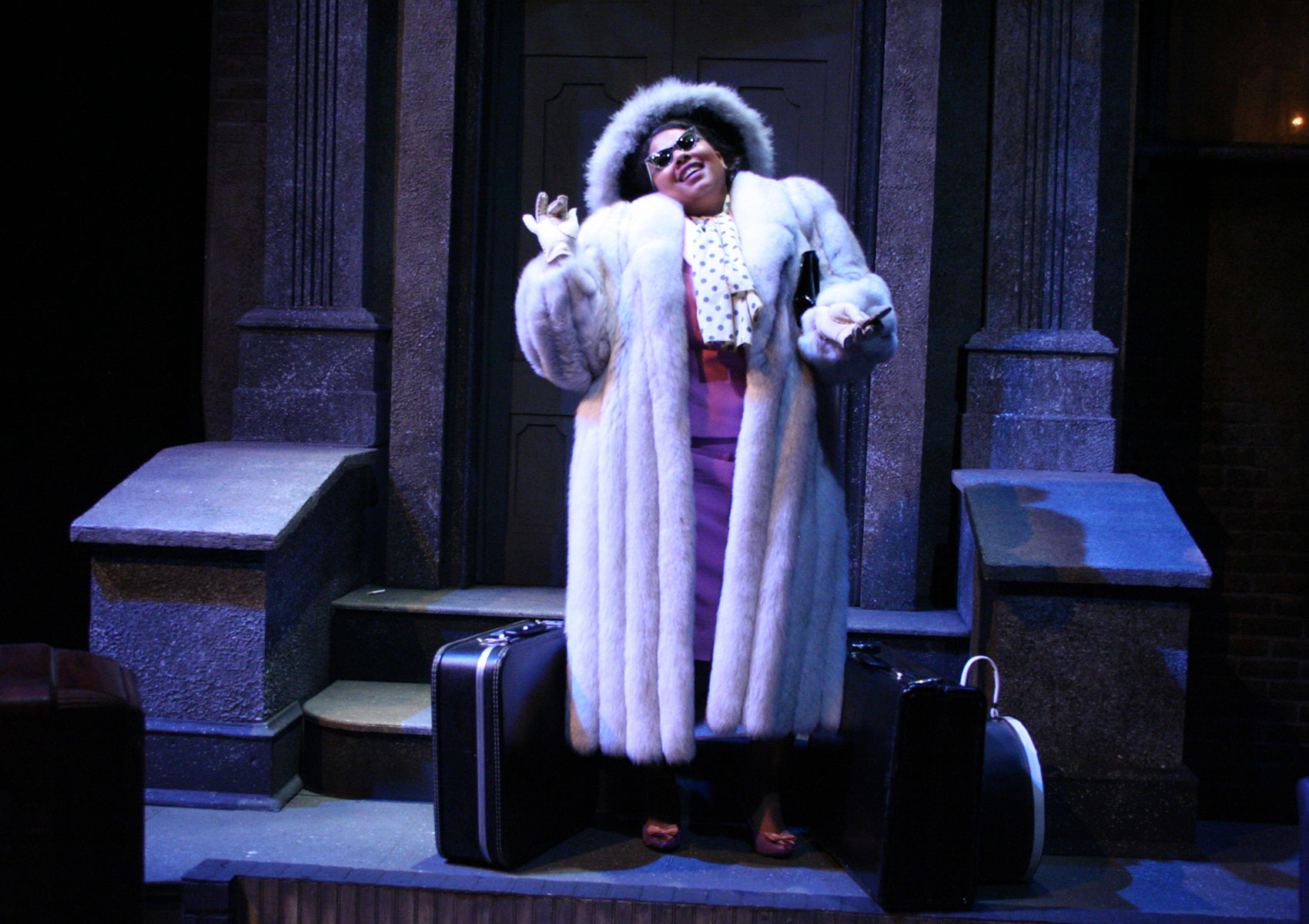 A woman in a fur coat and hat stands on steps with suitcases on either side of her in front of a door.