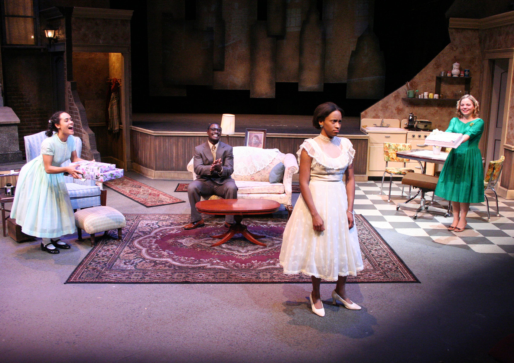 A woman stands frowning toward the left side of the audience while three characters smile in different places onstage behind her. Behind her toward the stage right a woman stands clapping, a man closer behind the girl claps while sitting on a couch and a woman holds a cake back toward stage left.