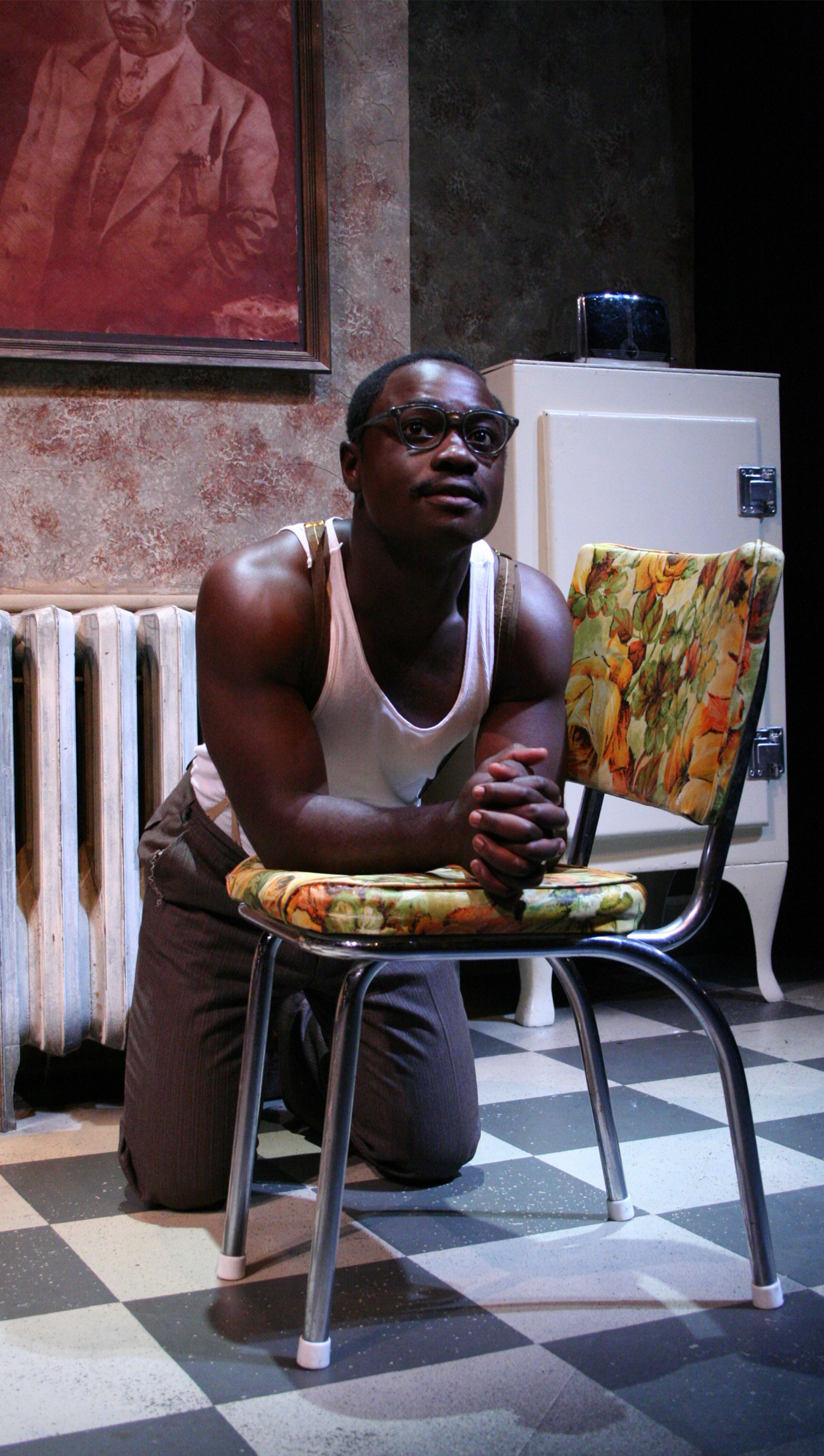 A man is on his knees leaning on a chair, his arms flat against the cushion and his hands together with the fingers interlocked. He is looking up and out toward the audience.