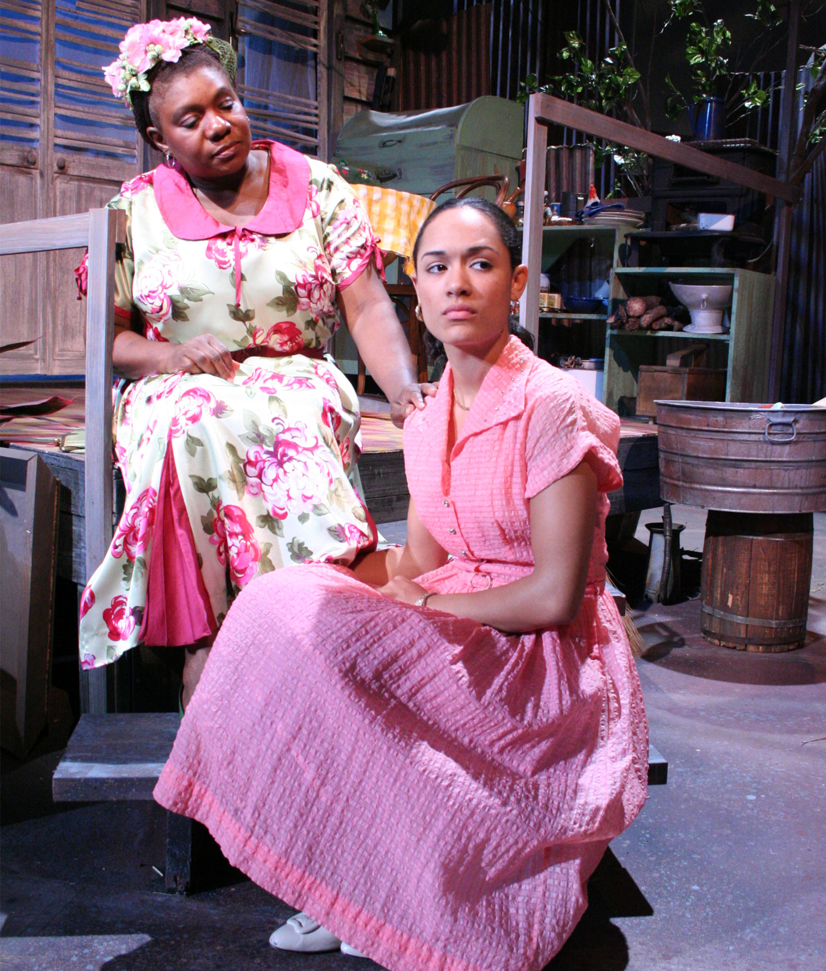 Two women sit together on porch steps. The older woman of the pair looks down at the younger woman as she sits on the step above the young woman. The older woman has one hand resting on the young woman’s shoulder while she has the other resting in her lap. The young woman sits upright and looks sadly down and outward toward stage left. 