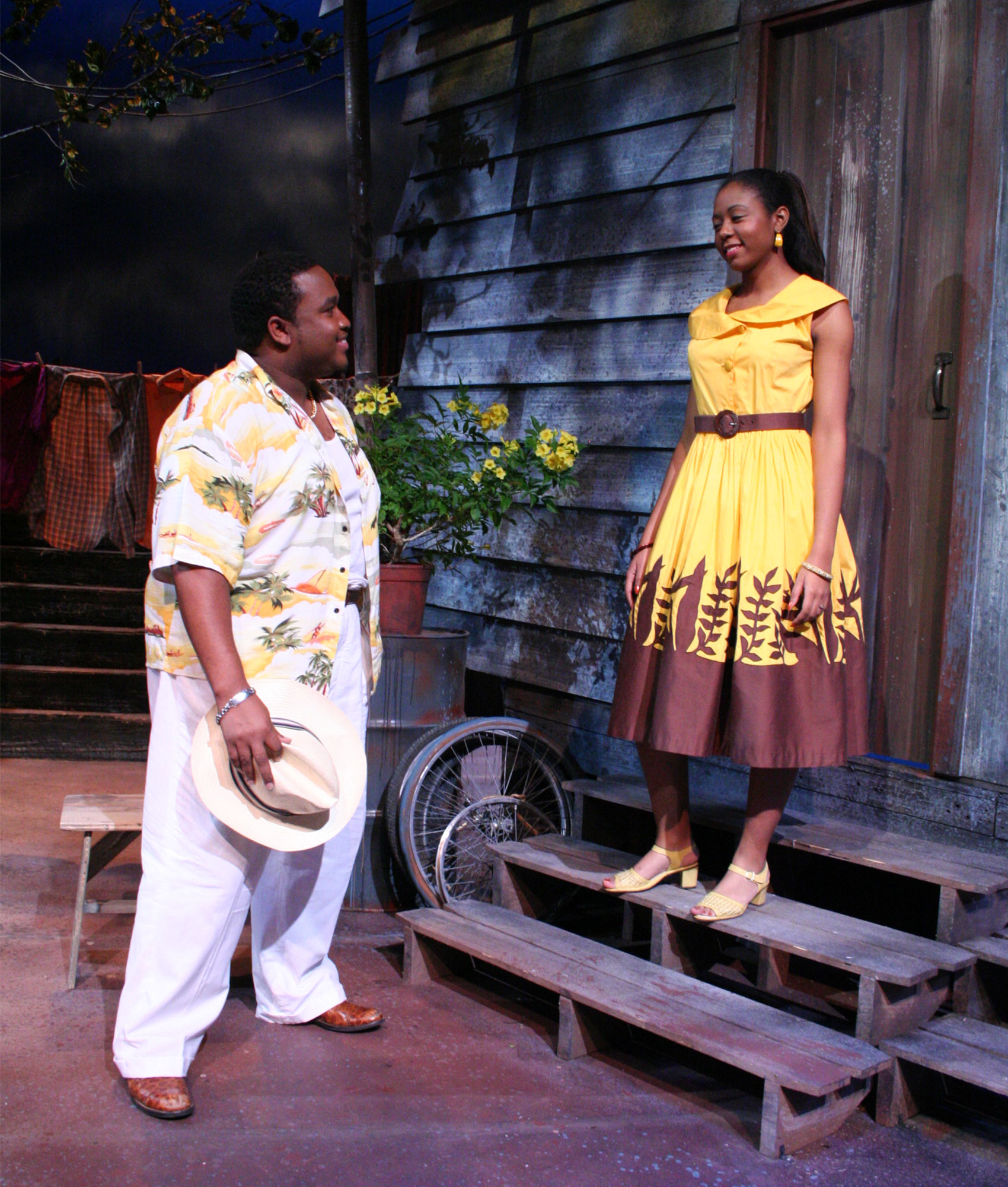 A man holds a hat in his hand at his side as he smiles at a woman standing on porch steps. The woman is smiling back down at the man. 