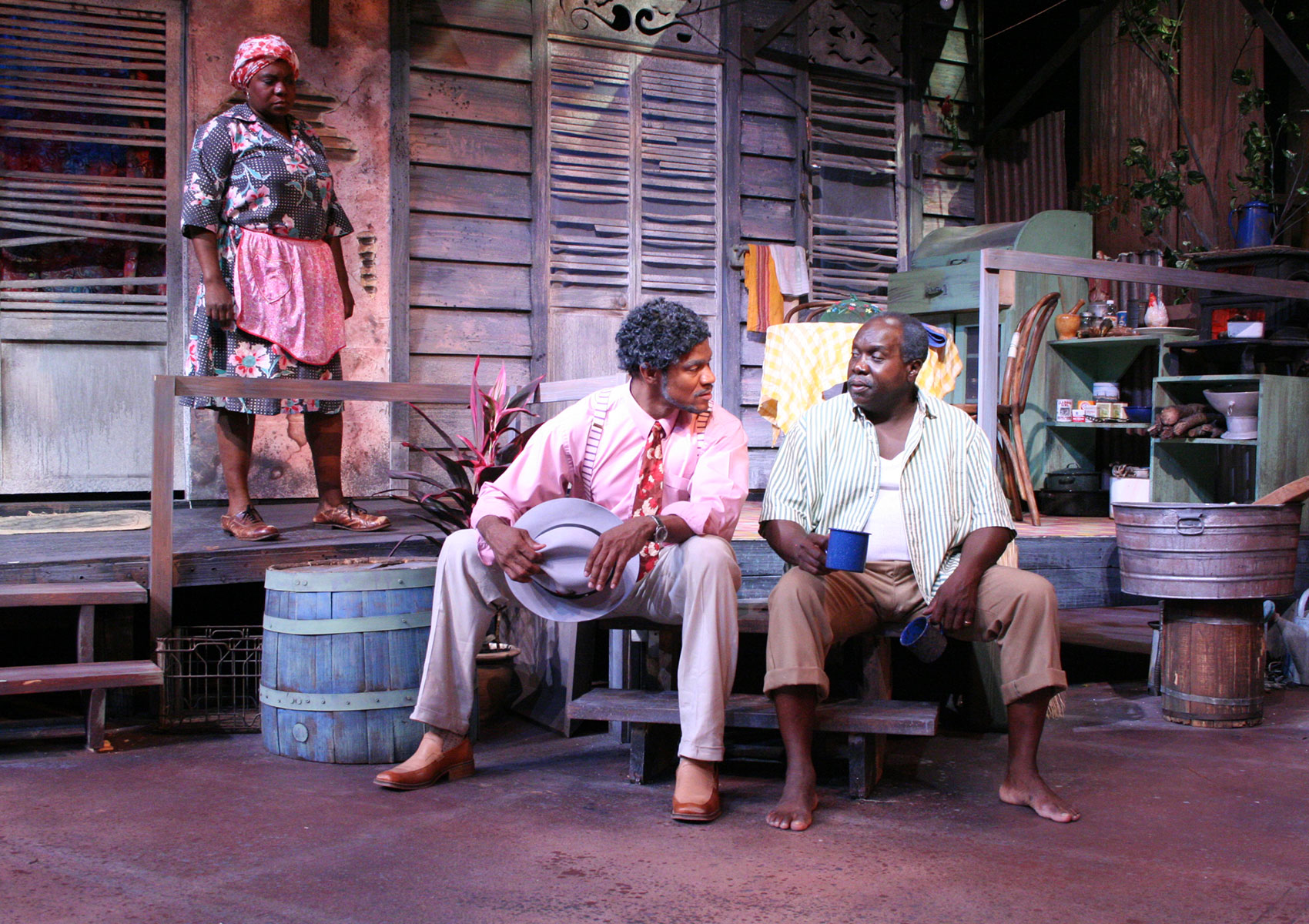 Two men sit on porch steps with their legs slightly spread as they are positioned in conversation with each other. The man on the left holds a hat in both hand and is leaning toward the man on the right, who is holding a coffee mug in each hand (one arm slackened while the other is being held as if ready the mug is about to be drank from) and is looking as if he is speaking. An older woman is watching them with her brow slightly furrowed from on the porch. 