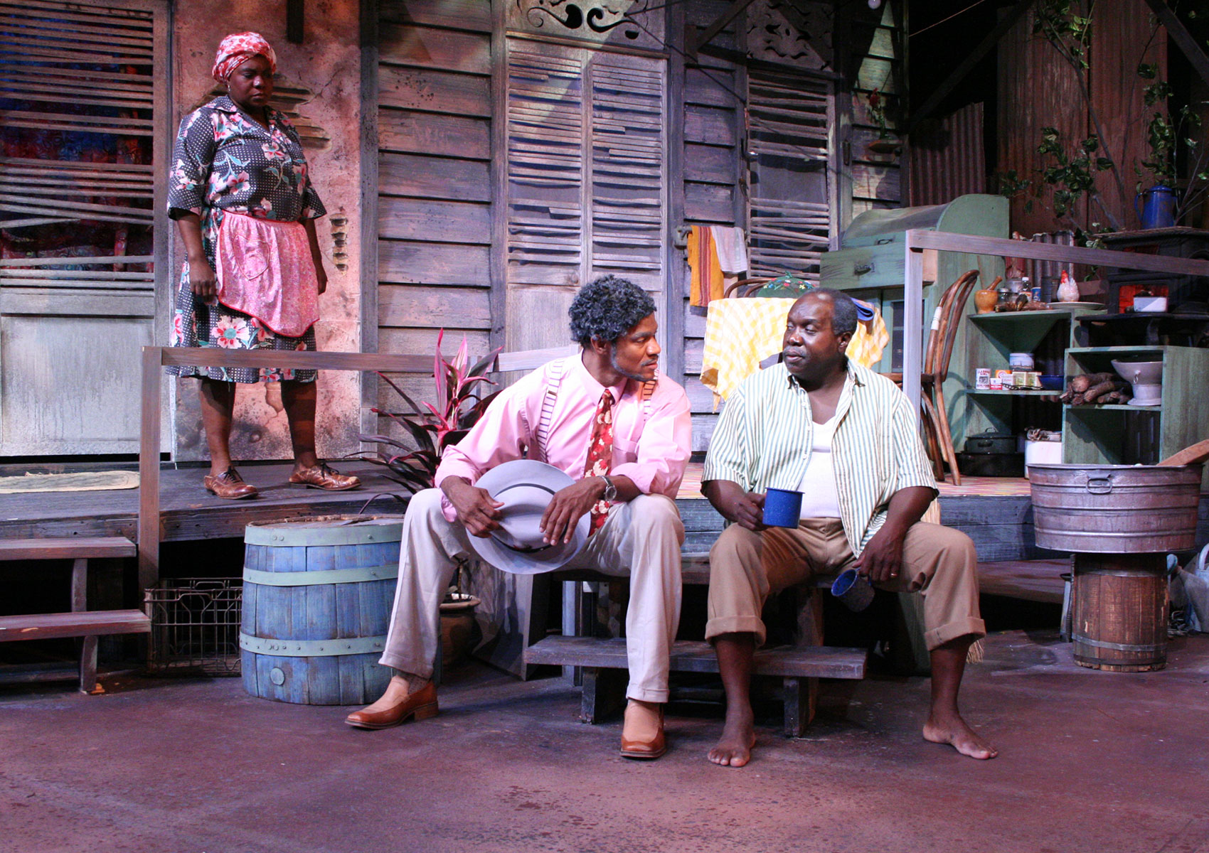Two men sit on porch steps with their legs slightly spread as they are positioned in conversation with each other. The man on the left holds a hat in both hand and is leaning toward the man on the right, who is holding a coffee mug in each hand (one arm slackened while the other is being held as if ready the mug is about to be drank from) and is looking as if he is speaking. An older woman is watching them with her brow slightly furrowed from on the porch.