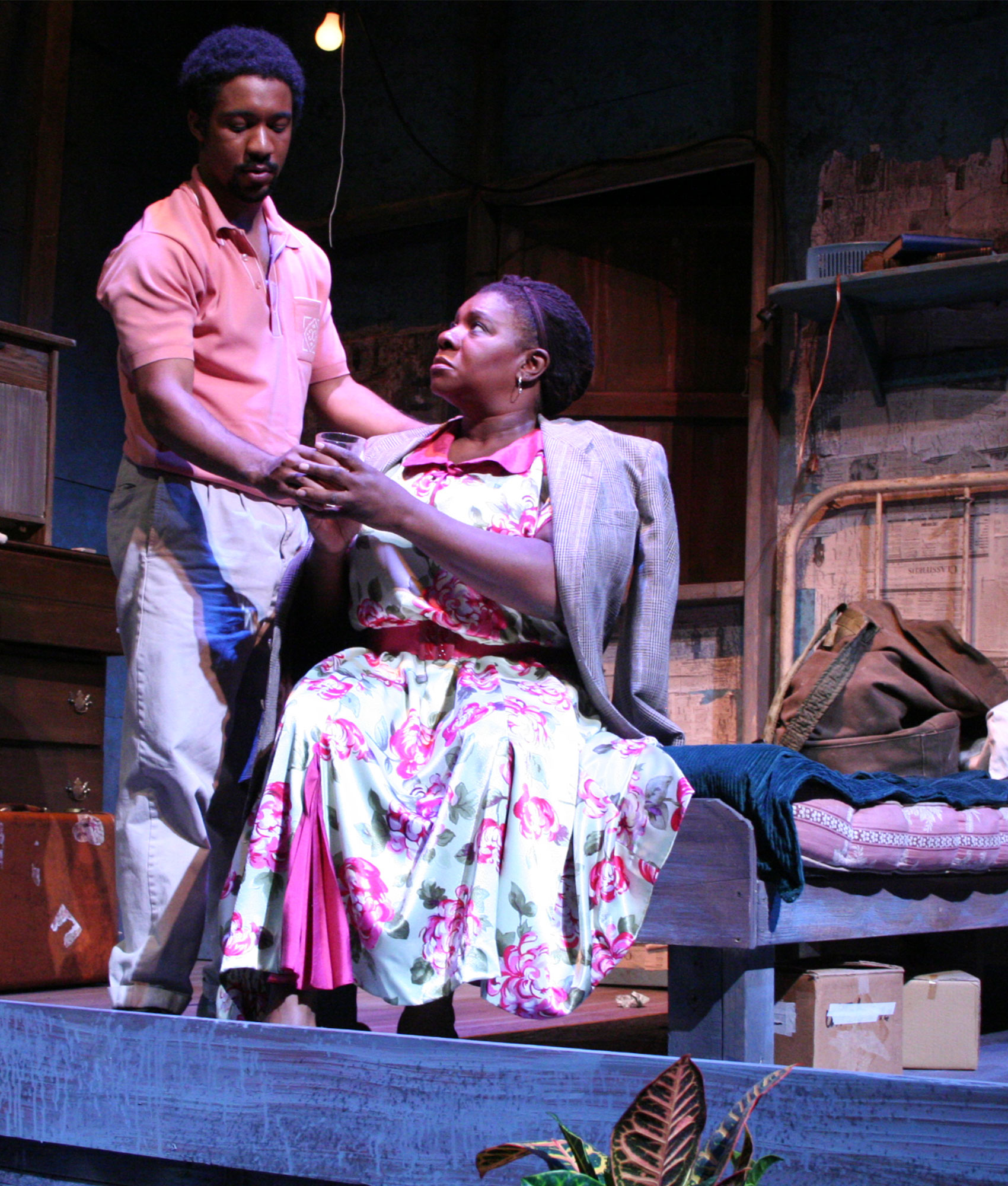 A young man stands above an older woman who sits on the edge of a bed looking up at the man. He is looking down away from the woman as he hands her a glass and has his hand resting on the back of her shoulder. The older woman holds the glass with both hands as she looks up at the young man, she has a suit jacket draped over her shoulders. 