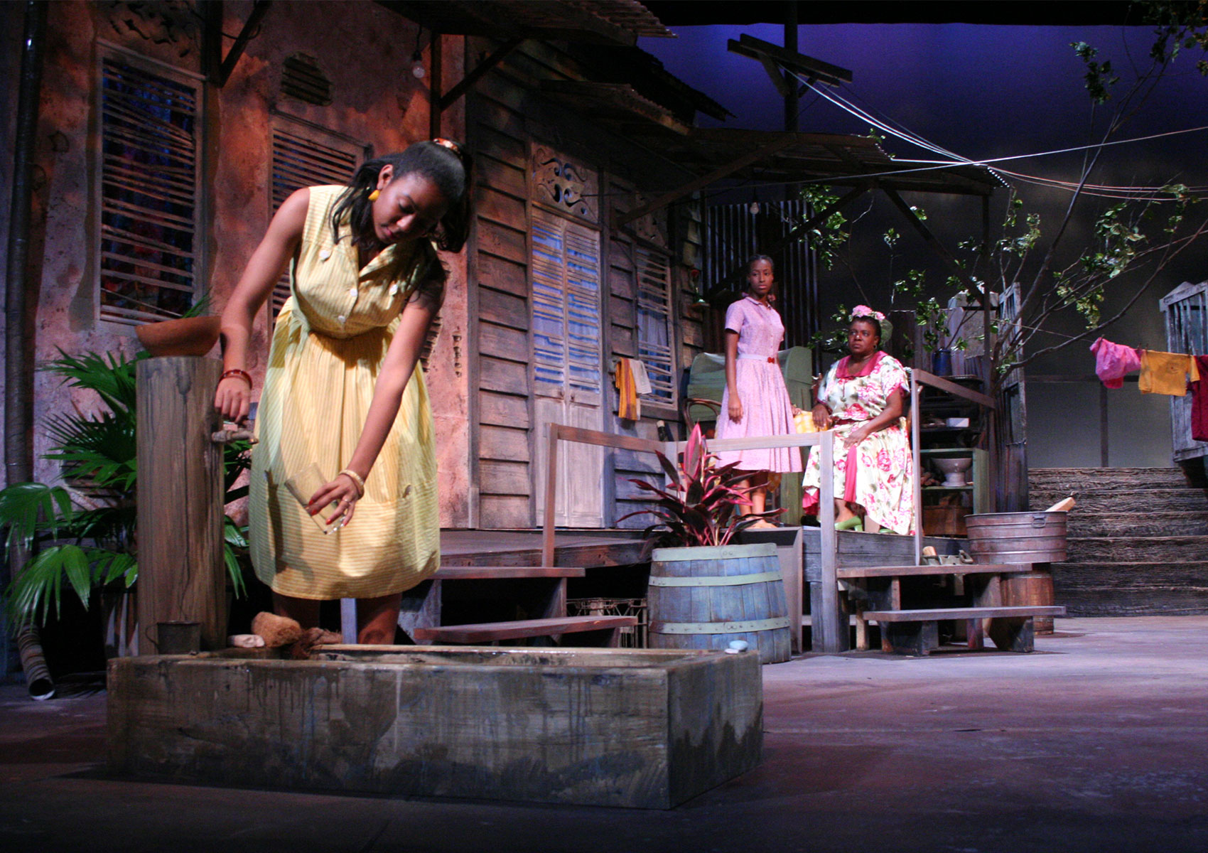 A young woman and an older woman stand and sit on a porch, respectively, as they watch a younger woman further downstage drawing water from a hose tap into a glass. 