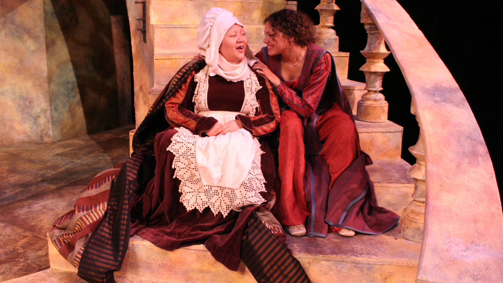 A woman and a nurse sit on a staircase, the woman facing the nurse and smiling while touching her shoulder. The nurse smiles back at the woman.