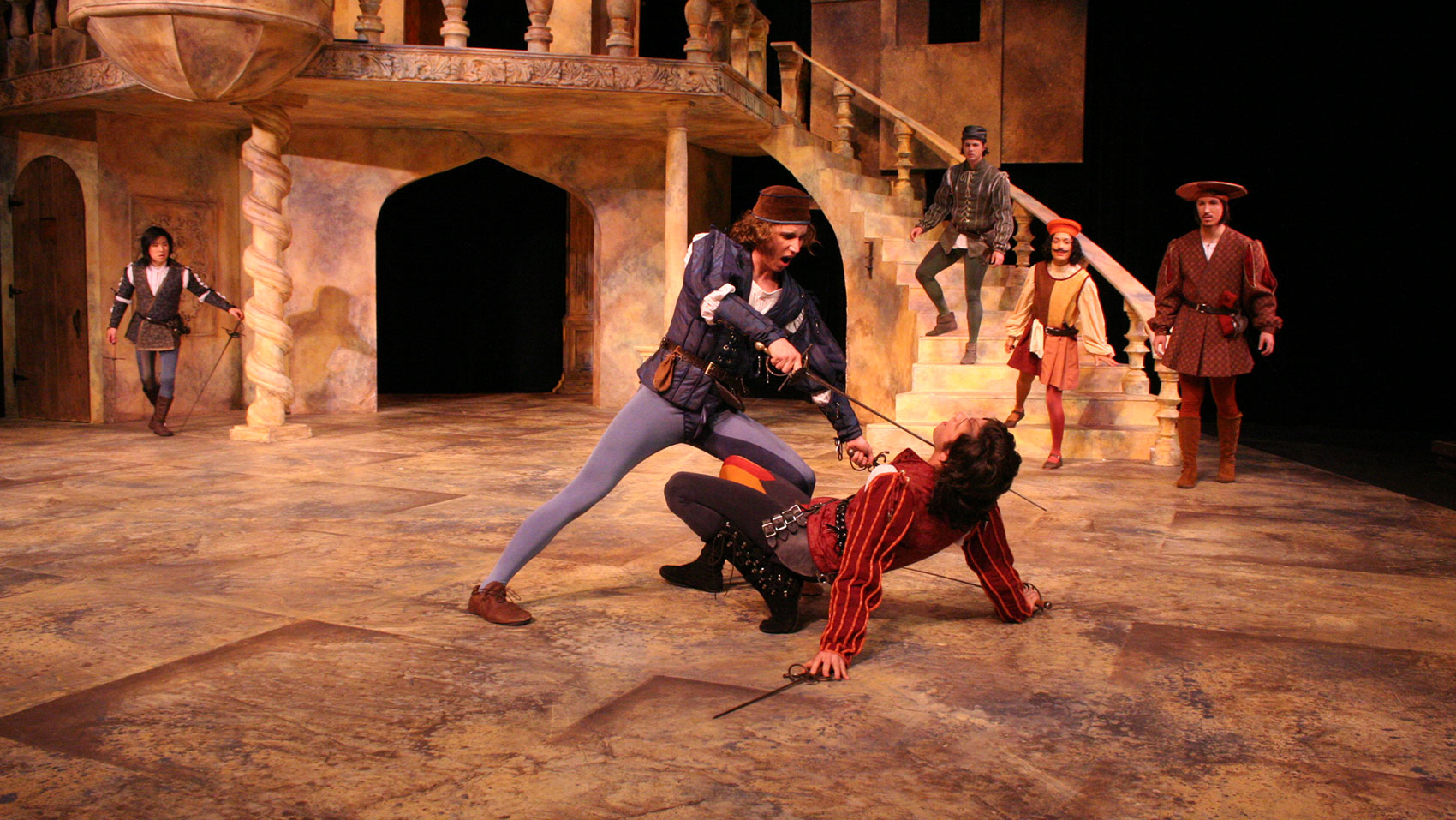 A man stands over a man, who is on the ground, with a sword pressed against his throat. Fpur characters watch from further back on the stage.