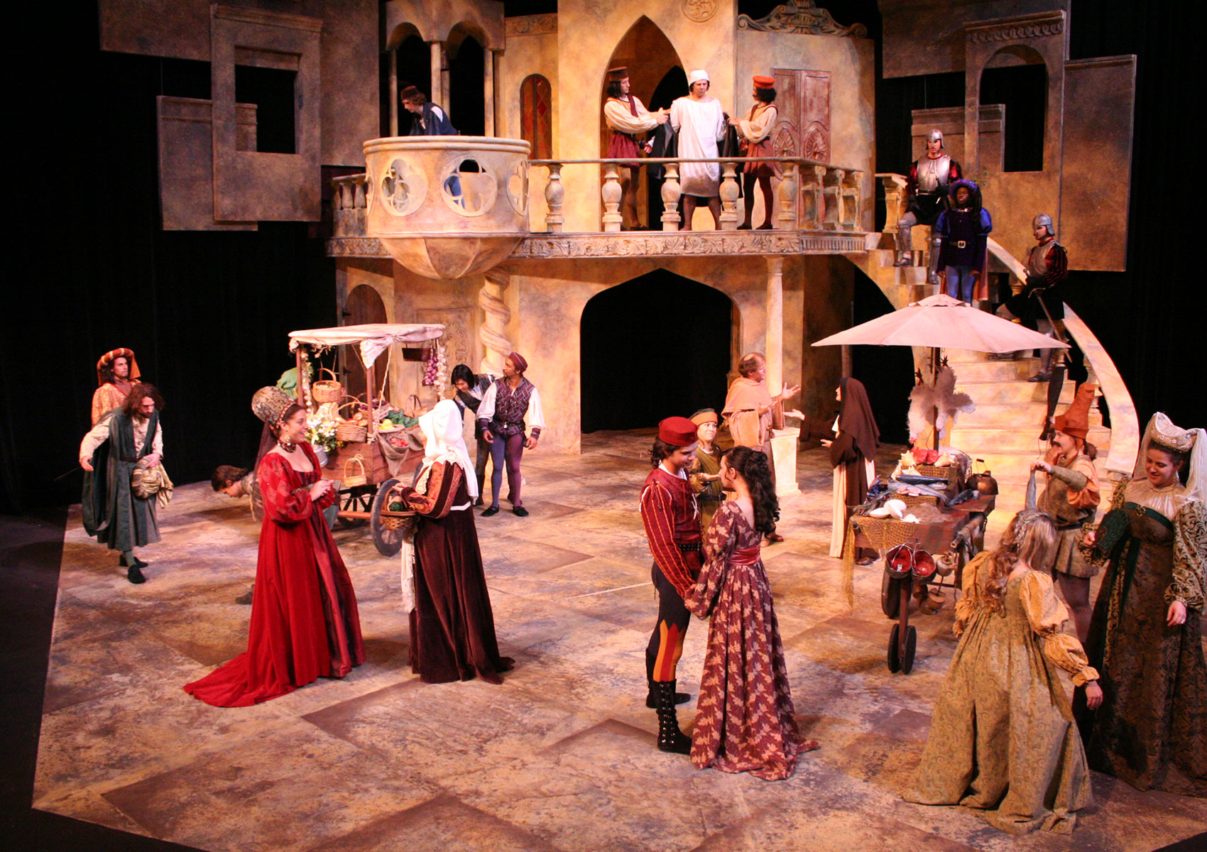 The stage is set like a market square. Carts of various wares are onstage, and characters are doing various actions: some are in conversation with each other, some are looking around, some are walking, and on a balcony two men hold a man by each of his arms.