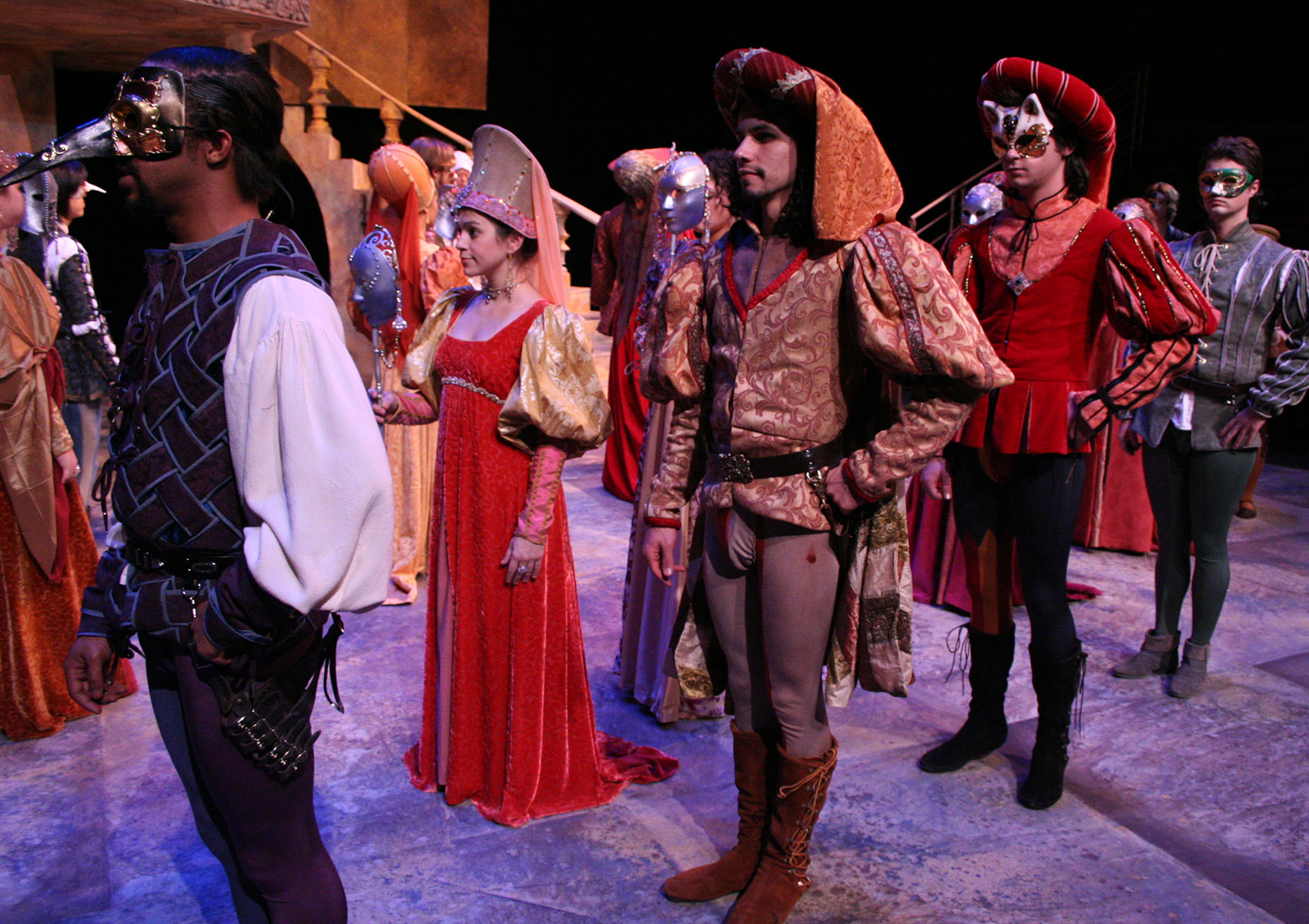 Many characters stand on stage in costumes, some wearing masks, in rows.
