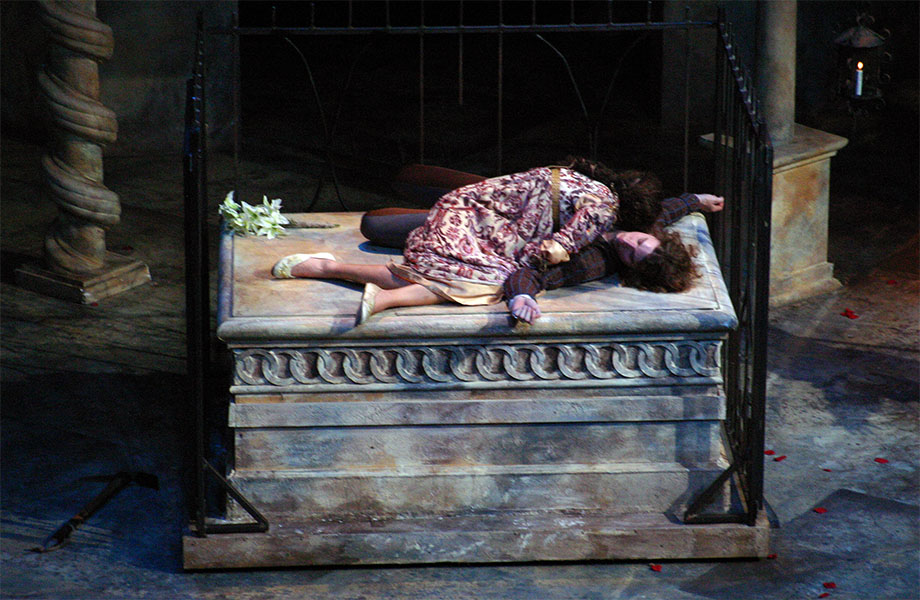 A woman sobs with her face in a male character’s chest while they are on top of a tomb with lilies.