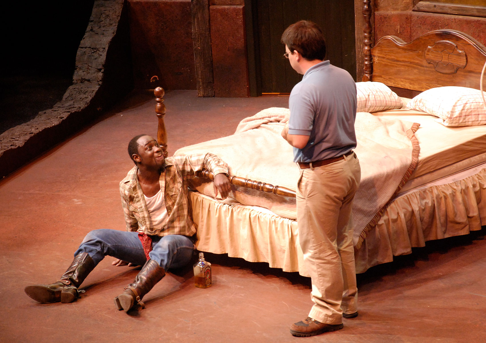 A man is on the floor handcuffed to a bedframe with a bottle of liquor next to him. He is looking up speaking animatedly to another man. 