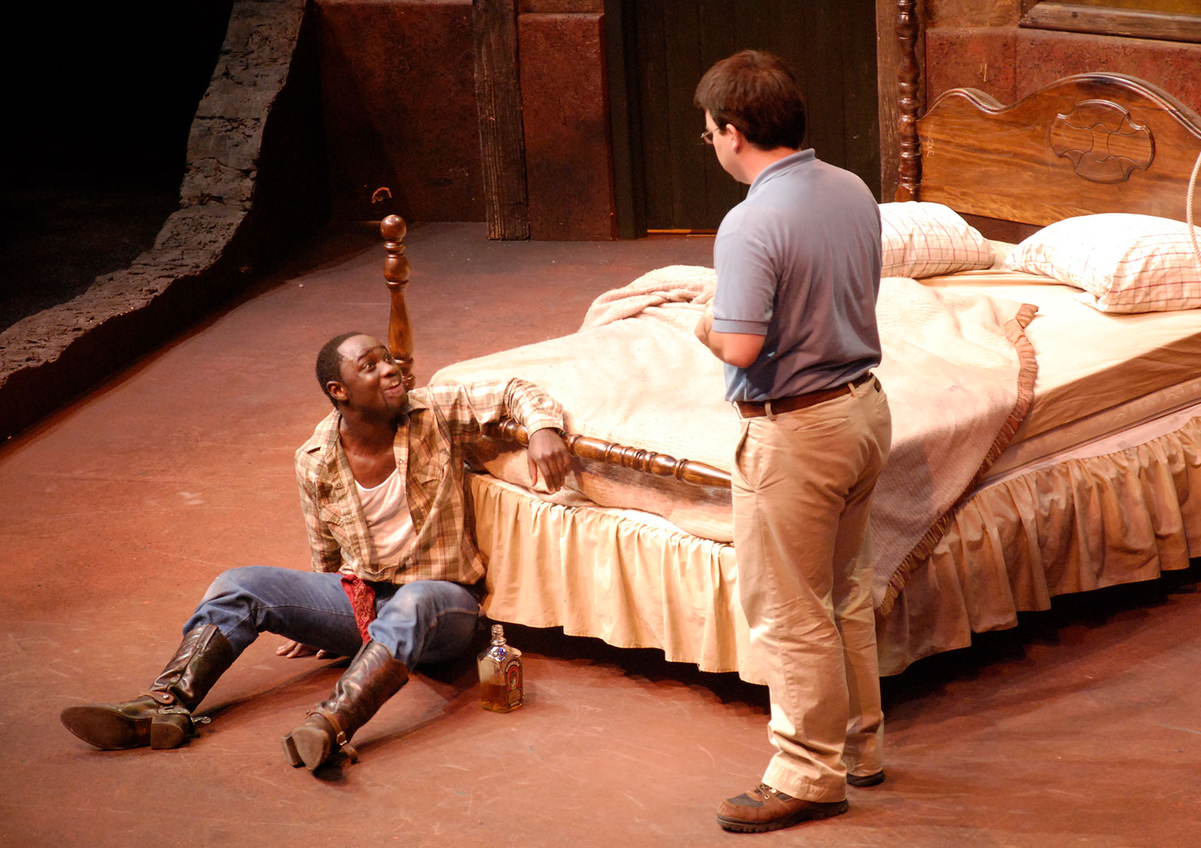 A man is on the floor handcuffed to a bedframe with a bottle of liquor next to him. He is looking up speaking animatedly to another man.