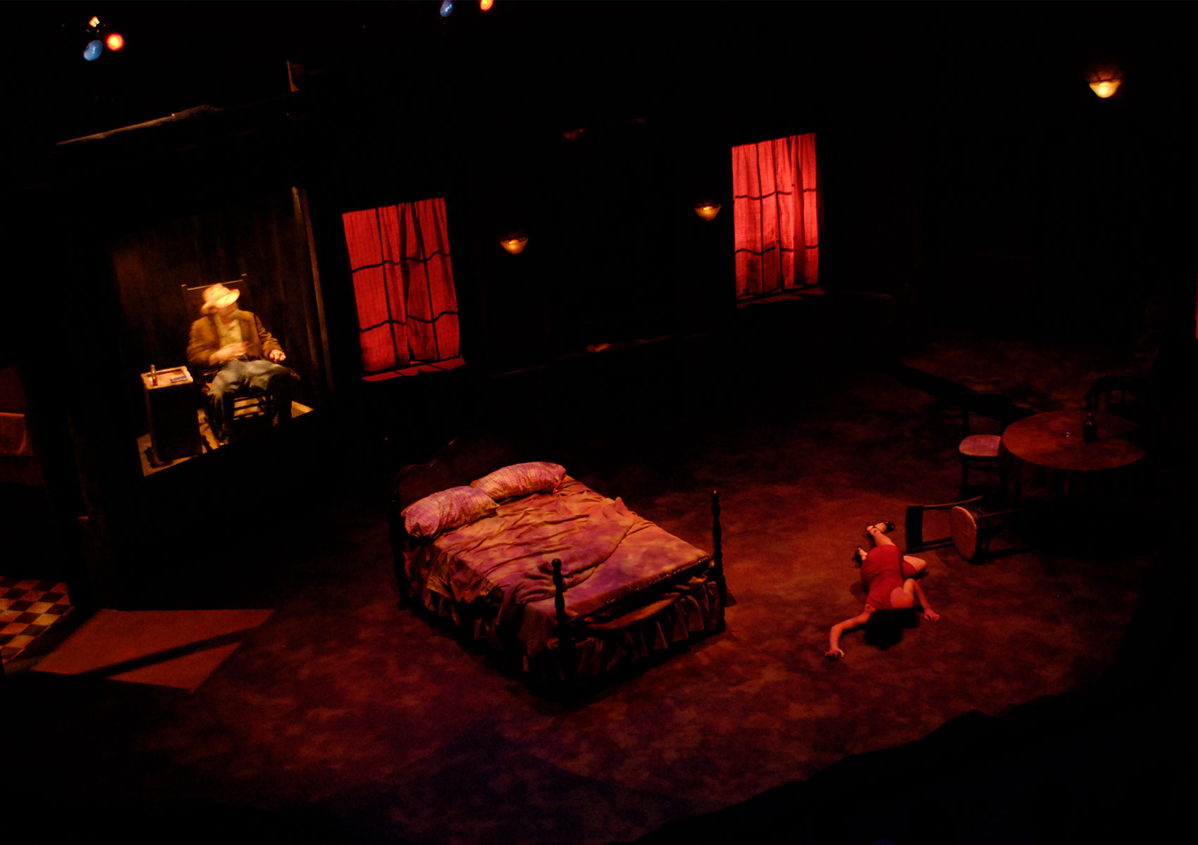 A woman lays sprawled on her stomach on the floor of a bedroom, the whole stage is flooded with red light. The older man in a cowboy hat is spotlighted in yellow light while sitting in a rocking chair next to a box.