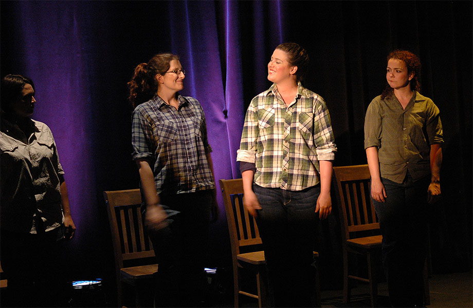 A group of four women are on stage in front of chairs, the two spotlighted in the center are smiling at each other.