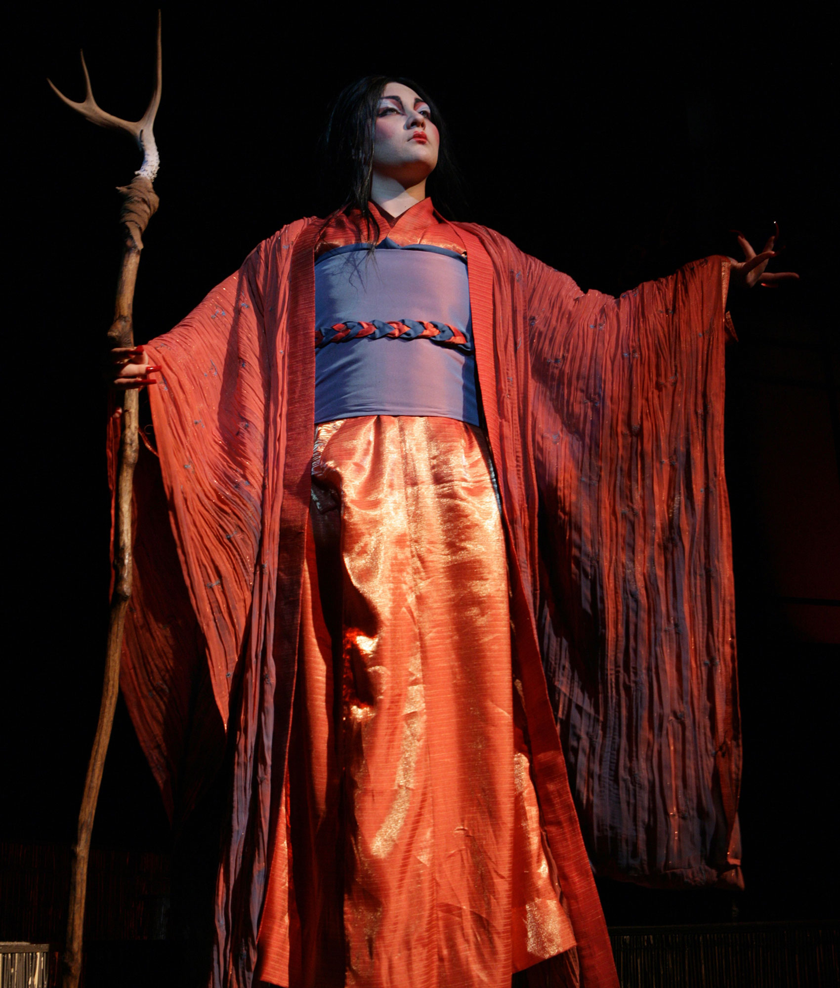 A woman with colorful makeup and attire holds a wooden staff topped with an antler in one hand and raises her other palm with her fingers outstretched.