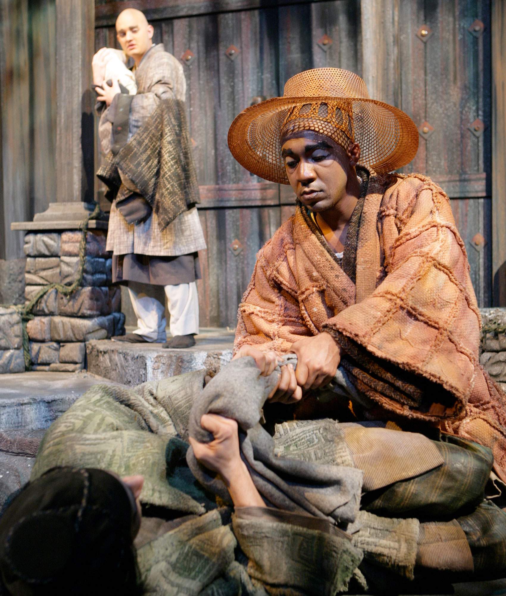 A man in orange, cloth clothing and a hat holds on one end of a cloth with both hands tightly while kneeling on the ground. A woman lying on the ground holds the other part of the cloth with both hands, one nearer o the first person’s hands, as she looks at the person kneeling in front of her.