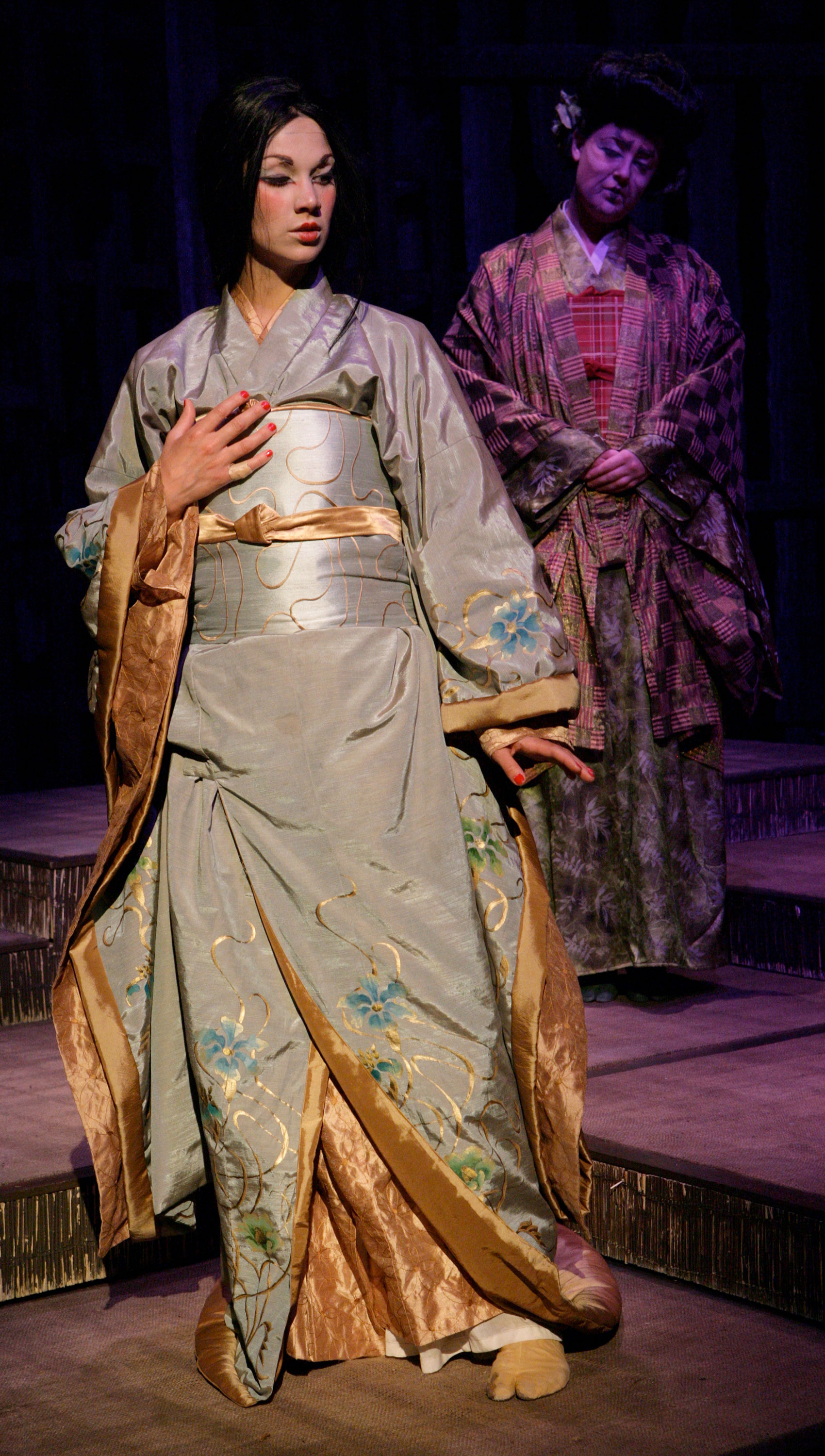 A woman in formal, floral attire accented in gold and makeup is spotlighted on stage. She is looking downward diagonal of stage left and has her right hand on her chest and her left hand at her side with the palm parallel to the floor. An older woman in multi-patterned attire watches the woman behind her from out of the spotlight. 